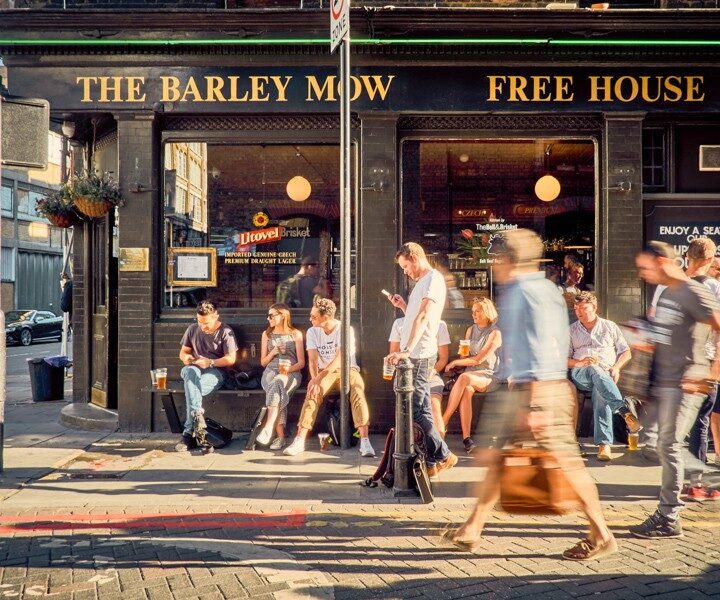 Exterior shot of The Barley Mow in Shoreditch, East London. People are sat on benches chatting whilst people walk past in the sun.